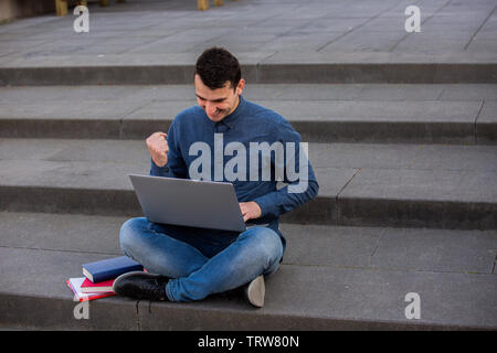 Students celebrating success using his laptop on capmus stairs. Happy Students winning with his project idea. Partnership concept. Students Teamwork. Stock Photo