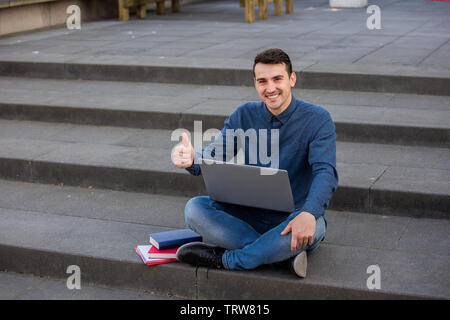 Smiling University student with thumb up gesture outdoors on Campus sitting on stairs using his laptop. A confident student navigates on internet with Stock Photo