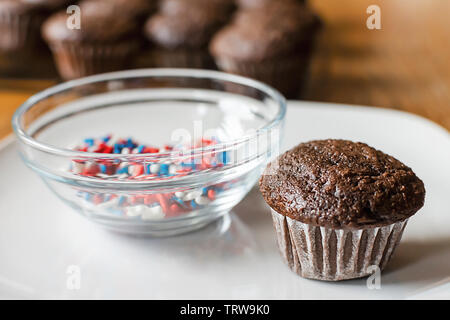 Chocolate mini cupcake on white plate waiting to be frosted.  Bowl of red, white, and blue sprinkles nearby.  Additional blurred cupcakes in the back Stock Photo