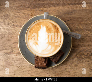 A cup of flat white coffee with chocolate brownies and a spoon on a saucer against a wood effect background, England, UK Stock Photo