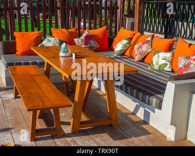 Dining table with wooden long chair and orange pillows on modern sofa on wooden floor on sunny day. Stock Photo