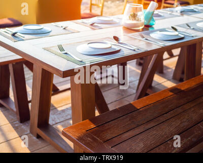 Plates, spoons, forks and knives on wooden table with wooden chairs on wooden floor near the beach. Table set on outdoor restaurant. Stock Photo