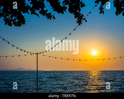 Light bulbs hanging on string wire and the sun on sunset sky and seascape background. Stock Photo