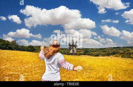 Beautiful Ukrainian girl in white ethnic embroider shirt running to the old wooden wind mill in National architecture museum in Pirogovo, Kiev, Ukrain Stock Photo