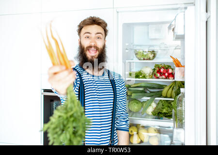Portrait of a well-looking man standing with carrot near the fridge full of fresh vegetables and fruits at home Stock Photo