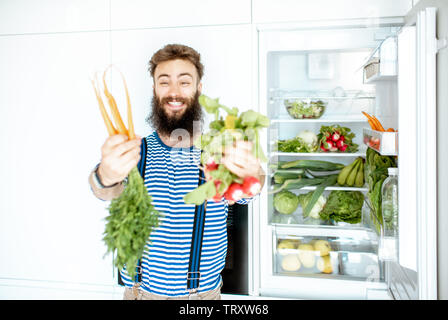 Portrait of a well-looking man standing with carrot and radish near the fridge full of fresh vegetables and fruits at home Stock Photo