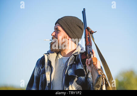 Man brutal bearded guy gamekeeper blue sky background. Hunter with rifle gun close up. Guy bearded hunter spend leisure hunting and smoking. Brutality and masculinity. Hunting masculine hobby concept. Stock Photo