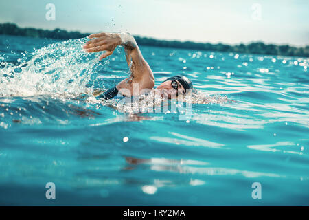 Professional triathlete swimming in river's open water. Man wearing swim equipment practicing triathlon on the beach in summer's day. Concept of healthy lifestyle, sport, action, motion and movement. Stock Photo