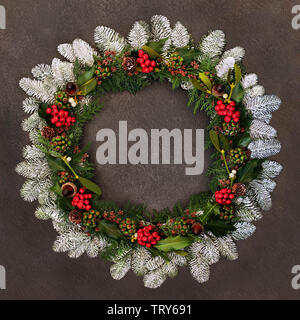 Natural winter and Christmas wreath with holly, ivy, mistletoe, snow covered fir, acorns, pine cones and cedar leaves on grunge background. Stock Photo