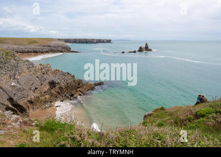 A photographer sitting on the cliff overlooking rugged coastal scenery at Broad Haven in Pembrokeshire, Wales, with Church Rock in the sea Stock Photo