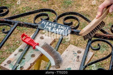 Man’s hand holding a wire brush to remove black paint from a rusty ornate metal gate, lying on a workbench and bearing a sign ‘please shut the gate’. Stock Photo