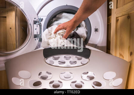 Man’s hand putting dirty bedding into a washing machine, with a laundry basket in front of the open cupboard doors for the appliance. Stock Photo
