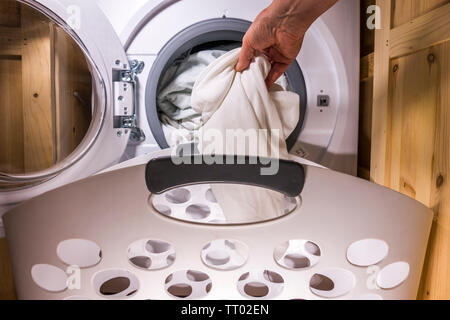 Man’s hand putting dirty bedding into a washing machine, with a laundry basket in front of the open cupboard doors for the appliance. Stock Photo