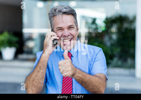 Mature businessman at phone making money and showing thumb up Stock Photo
