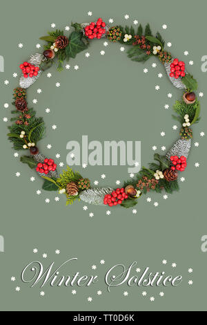 Natural winter solstice wreath with holly, snow covered fir, mistletoe, acorns, cedar leaves, ivy & loose snowflakes on green background. Stock Photo