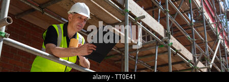 Panoramic web banner male builder foreman, construction worker, surveyor, engineer or architect on site holding a clipboard and drinking a mug of coff Stock Photo