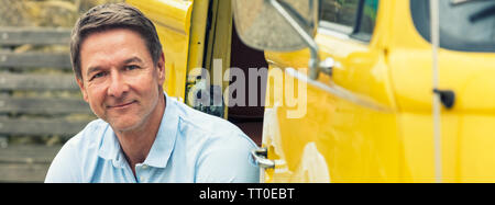 Panoramic web banner portrait shot of an attractive, successful and happy middle aged man male wearing a blue polo shirt, sitting smiling in the doorw Stock Photo