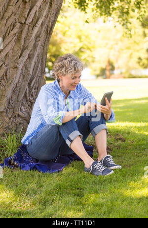 Happy, smiling, middle aged mature woman using a digital tablet computer e-reader outdoors in a park. She is touching the screen. Stock Photo