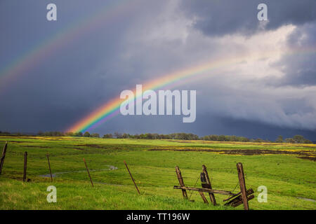 Amazing double rainbow over a tranquil meadow landscape bordered by an old broken barbed wire fence. Stock Photo
