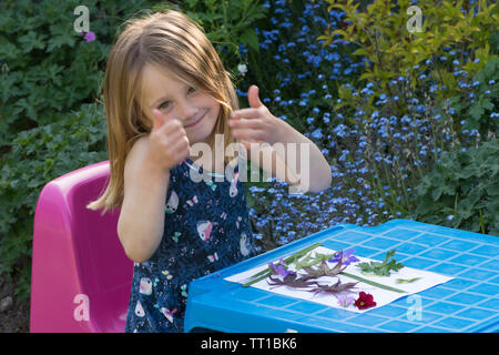 three year old girl giving thumbs up sitting at a small table, proud of a picture made from flowers and leaves stuck on paper, nature art craft Stock Photo