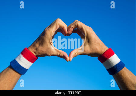 Proud American athlete making hand heart with USA colors red white and blue wristbands against bright sky copy space Stock Photo