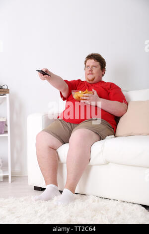 Lazy overweight male sitting on couch with chips and watching television Stock Photo