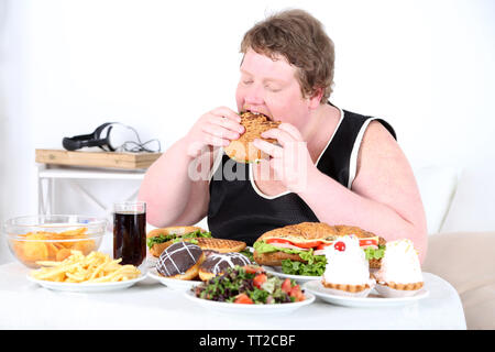 Fat man eating a lot of unhealthy food, on home interior background Stock Photo