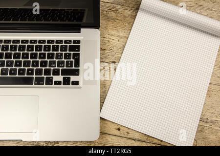 Contrast between old and new office work: shot from the top of an aluminum laptop on a wooden desk with an open checkered notebook with blank sheet Stock Photo