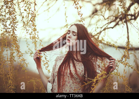 Portrait of a romantic girl with red long hair in the wind under a willow tree. Stock Photo