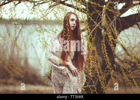 Portrait of a romantic girl with red long hair in the wind under a willow tree. Stock Photo