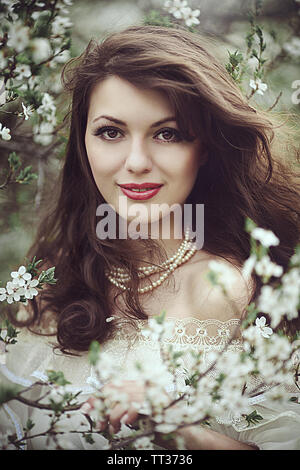 A beautiful young girl stands among the flowering trees. White flowers. Spring. A girl with her hair in a white dress. Portrait. Romance. Stock Photo