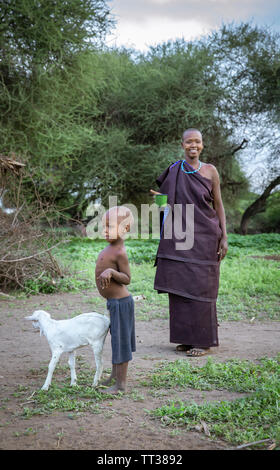 Same, Tanzania, 4th June, 2019:   Maasai  boy with a baby goat and mother smiling at the background Stock Photo