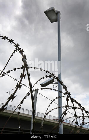 Close-up of razor wire and barbed wire surounding a secure compound in east London, UK. Shows security camera and lighting. Road viduct beyond. Stock Photo