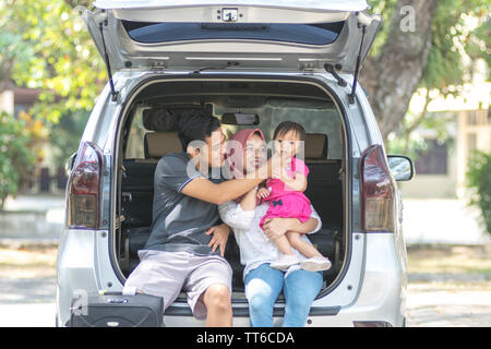 young muslim family , transport, leisure, road trip and people concept - happy man, woman and little girl sitting on trunk of hatchback car and laughi Stock Photo