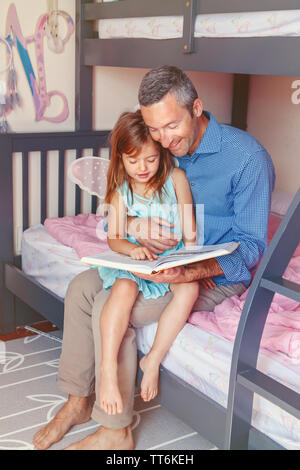 Family two people sitting on bed in bedroom reading book. Father and daughters girl at home spending time together. Parent talking communicate to chil Stock Photo