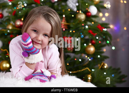 Little girl with mittens lying on fur carpet on Christmas tree background Stock Photo