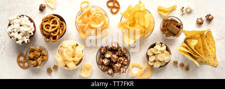 Assortment of Unhealthy Snacks: chips, popcorn, nachos, pretzels, onion rings in bowls, top view, banner. Unhealthy eating concept. Stock Photo