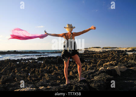 Isolated blonde woman with straw hat, batik dress, waving scarf in breeze standing on rocks at the ocean in evening sun - Fuerteventura, El Cotillo Stock Photo