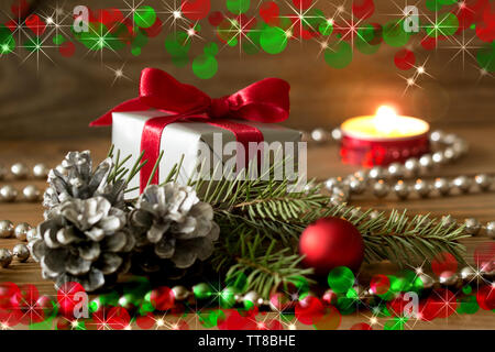 Red and green christmas composition: a gift wrapped with red ribbon, silver cones, red baubles and a fir tree branch - burning candle in background Stock Photo