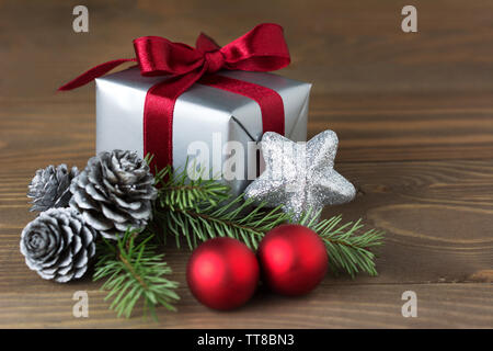 Christmas composition: a gift wrapped with red ribbon, silver cones, red baubles and a fir tree branch - isolated on wooden background Stock Photo