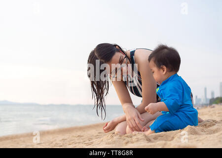 happy young mother smiling and looking at her little baby boy playing sand on tropical beach vacation at summertime. people family lifestyle and trave