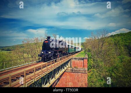 Beautiful old steam train driving along a bridge in the countryside. Concept for travel, transportation and retro old style. Stock Photo