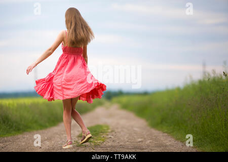 Back view of young romantic slim woman in red dress with long hair walking by ground road along green field on sunny summer day on blue sky copy space Stock Photo