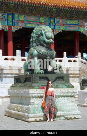 A white, caucasian, female western tourist, dressed fashionable and wearing sunglasses (shades) poses with a metal statue of a pouncing Chinese dragon Stock Photo