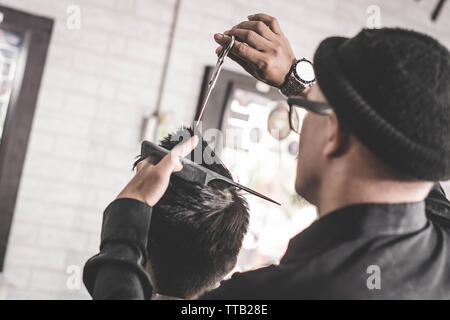 rear view of barber man with black shirt cutting his male costumer with scissor and comb in barbershop Stock Photo