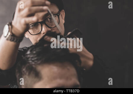 close up portrait of professional barber making hair cut of his attractive man costumer with scissors and comb in vintage barbershop Stock Photo