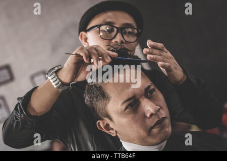 close up portrait of barber hairstyle styling hair of his costumer hair who sitting in chair with hand pomade and comb in barbershop or salon Stock Photo
