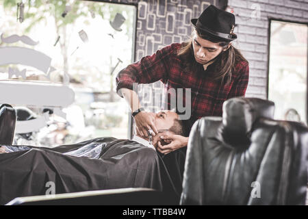 close up portrait of Asian barber with long hair shaving his costumer beard at barbershop chair while his male costumer relaxed at barber chair Stock Photo