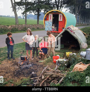 1960s, historical, Irish travellers... daytime and beside a country road in rural Ireland, a mother with her children cooking a pot on a fire outside  their home, a traditional barrel-roofed or bow top horse-drawn gypsy wagon and old tent. Stock Photo