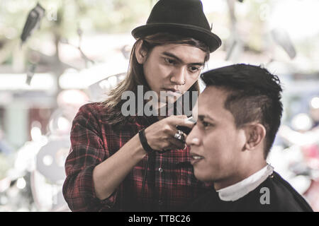 portrait of professional Asian man with long brown hair work as barber trimming his costumer with scissors and comb in barbershop Stock Photo